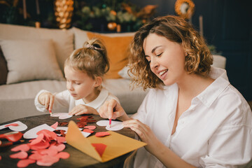 Mother and daughter making Valentine's day cards using color paper, scissors and pencil, sitting by...