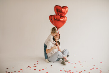 Young mother and daughter holding red heart shaped balloons on white background. Saint Valentine's...