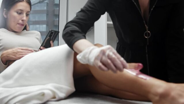 Waxing procedure - applying pink hot wax on the leg using a spatula while the woman sitting on a couch and looking at her phone