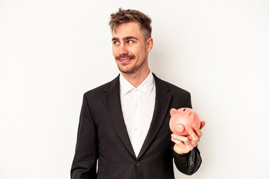 Young caucasian business man holding piggy bank isolated on white background looks aside smiling, cheerful and pleasant.