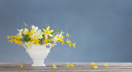 white and yellow spring flowers in vase on wooden table on blue background