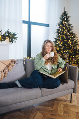 Young woman on the couch reads a book and enjoys coffee