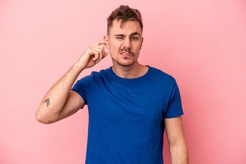 Obraz premium Young caucasian man isolated on pink background covering ears with fingers, stressed and desperate by a loudly ambient.