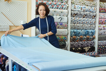 Beautiful Asian woman dressmaker unrolls a roll of blue fabric for cutting. Small business concept.