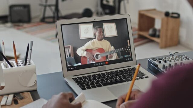 Over-shoulder of cropped student sitting at desk in his room in morning, taking notes when video calling via portable computer with Black man who playing guitar