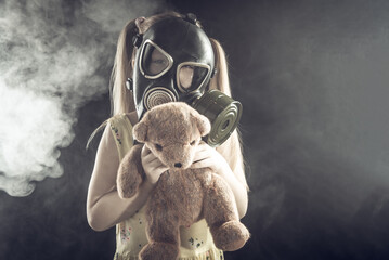 Little girl in a gas mask in smoke with a bear in her arms on a black background. Air pollution,...