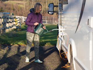 A lady motor homer uses a watering can to refill her recreational vehicle water tank through a side locker door.She wears camouflage trousers and flowery shoes.