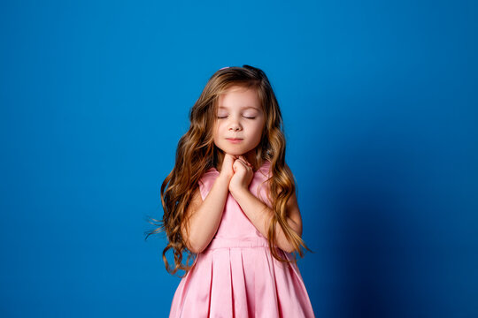 cute little girl with curls in a pink dress makes a wish and dreams standing with her eyes closed. studio background on a blue background. space for text. High quality photo - Powered by Adobe