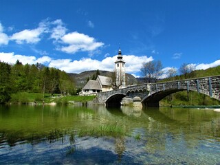 Beautiful view of the St. John the Baptist church next to Bohinj lake next to a arched stone bridge and a reflection in the lake and white clouds in blue sky