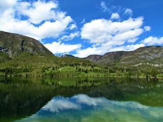 Fototapeta na wymiar Beautiful view of Bohinj lake in Gorenjska, Slovenia and a reflection in the lake and forest covered mountains and hill above and clouds in blue sky