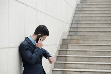 Young entrepreneur leaning on wall outdoors, talking on phone with colleague and checking time and notifications on smartwatch