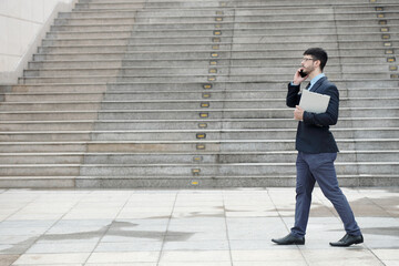 Side view of businessman walking outdoors, carrying documents folder and talking on phone with coworker