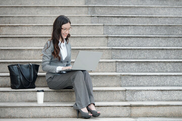 Smiling elegant businesswoman sitting on steps and working on laptop, checking documents or answering e-mails