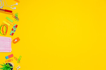 Series of stationery items on left of yellow background, colored pens, labels, paper clips, calculator, face mask and more back to school concept, flat lay, empty space