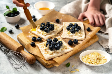 Raw puff pastry with cheese cream and blueberries. The process of making delicious sweet buns or mini pies. Greasing the edges of the baking dough with egg. Hands in the frame