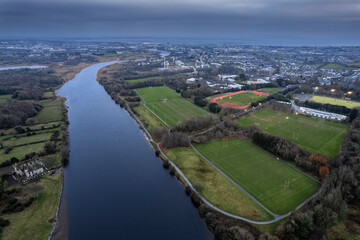 Fototapeta premium River Corrib flows into Galway city, aerial view. Sport ground with tall goal posts for Irish national sport on the right. Training field for hurling, rugby, camogie, Gaelic football. Dusk time.