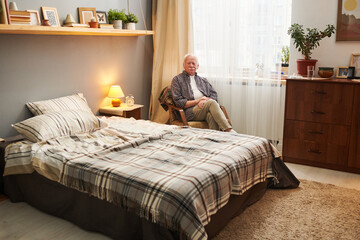 Portrait of senior man sitting on armchair and looking at camera, he resting in his bedroom