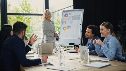 man pointing with hand while talking to middle aged businesswoman near smiling interracial colleagues