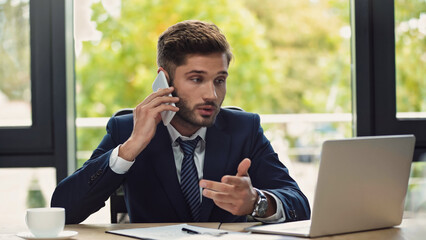 young businessman looking at laptop while talking on smartphone in office