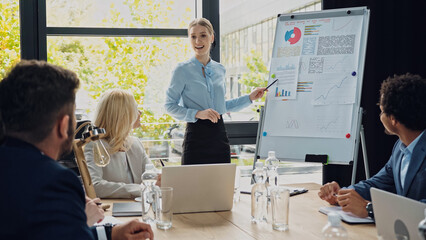 positive businesswoman pointing at flip chart and talking to interracial colleagues in meeting room