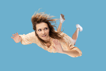 Beautiful young woman levitating in mid-air, falling down and her hair messed up soaring from wind, model flying hovering with dreamy peaceful expression. indoor shot isolated on blue background - obrazy, fototapety, plakaty