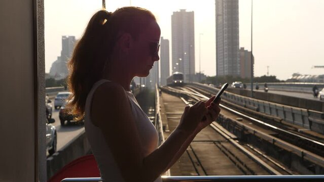 Woman standing with phone, subway line and road seen blurred in background, slow motion shot. Tourist lady using mobile application, stay at skytrain station. Concept of public transport at Bangkok