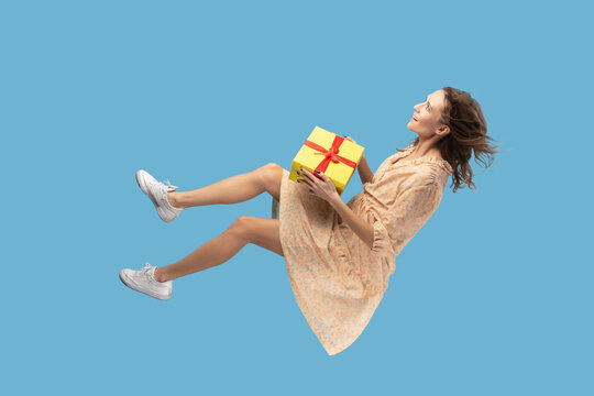 Hovering in air. Smiling happy woman in yellow dress levitating and holding her gift box, feeling joyful with her amazing birthday or christmas gift. indoor shot isolated on blue background.