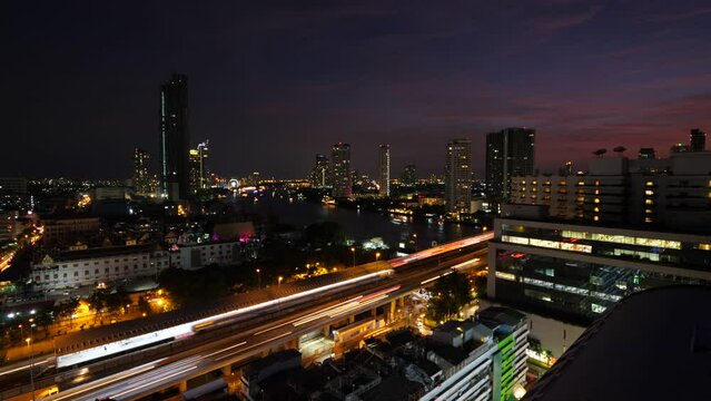 Lively flashing traffic on Sathon Road near King Taksin Bridge, night time lapse shot of Bangkok. Long lights from moving cars on roadway, urban view from high point. Boats move on Chaophraya river