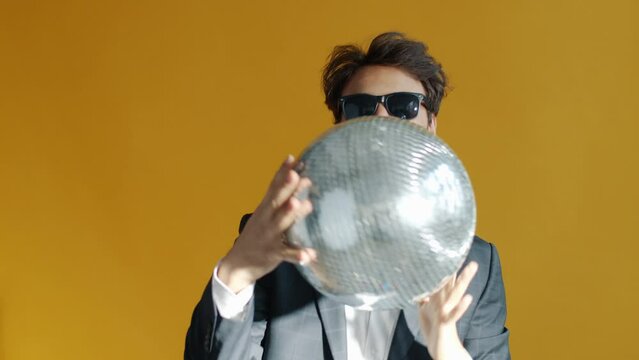 Slow motion portrait of cheerful mixed race man holding glitterball and dancing on yellow background. Celebration and happiness concept.