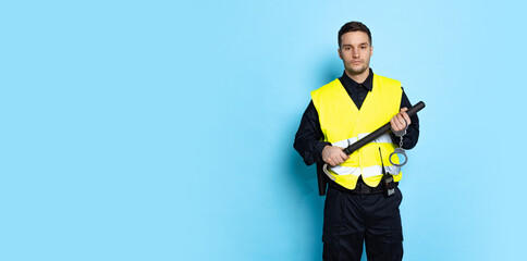 Half-length portrait of young male policeman officer wearing black uniform and warnvest posing isolated on blue background. Concept of job, caree, law and order.