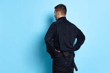 Obraz na płótnie Canvas Back view of young man, policeman officer wearing black uniform isolated on blue background. Concept of job, caree, law and order.