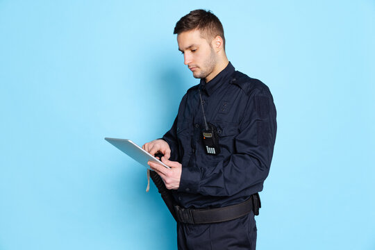 Portrait of young man, policeman officer wearing black uniform using tablet isolated on blue background. Concept of job, caree, law and order.