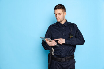 Portrait of young man, policeman officer wearing black uniform using tablet isolated on blue background. Concept of job, caree, law and order.