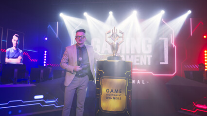 Man host with microphone walking and talking to audience while demonstrating golden cup on stand during esports championship - obrazy, fototapety, plakaty