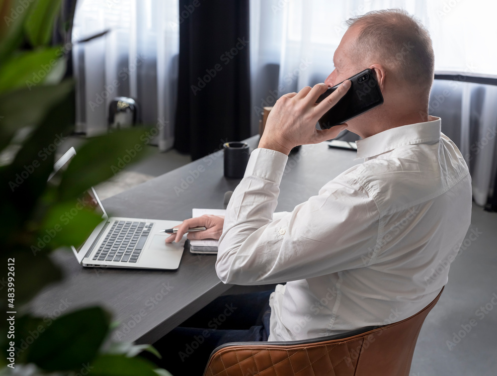 Poster mature businessman sitting at table in office, talking on mobile phone while looking at laptop scree