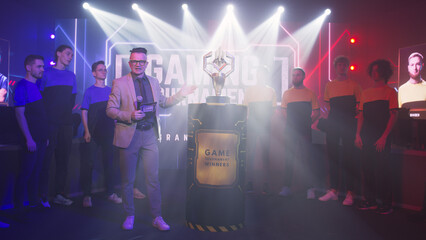 Host man speaking about golden cup while standing near cyber esportsmen at start of gaming tournament - obrazy, fototapety, plakaty