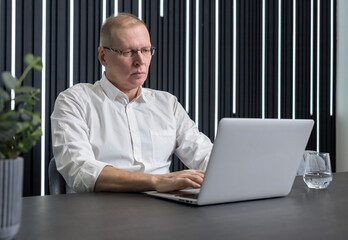 Concentrated and serious mature businessman in glasses sitting at table and typing on laptop keyboard. Employee working in office. High quality photo