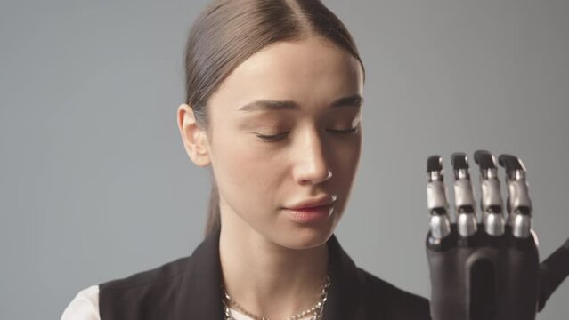 Slowmo close-up portrait of smiling young Caucasian woman with black bionic arm looking at camera on gray background