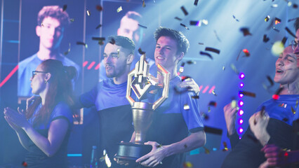 Happy professional esport team of gamers winners with golden cup standing under falling confetti and celebrating victory in professional esports gaming tournament together © Framestock