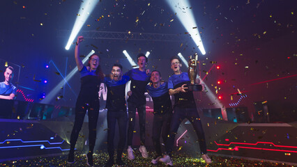 Professional cybersport team of gamers winners with golden trophy hugging and jumping amidst confetti and pyrotechnics while celebrating victory in esports competition © Framestock
