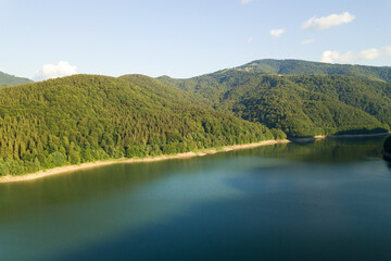 Obraz premium Aerial view of big lake with clear blue water between high mountain hills covered with dense evergreen forest
