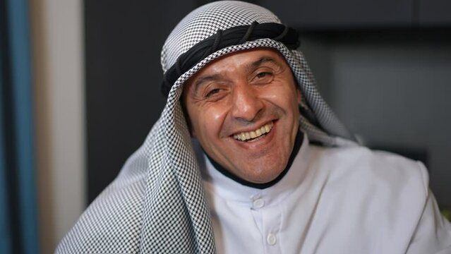 Front view portrait of cheerful Middle Eastern handsome man in traditional dishdasha and chequered keffiyeh laughing out loud looking at camera. Positive joyful guy posing indoors in kitchen at home