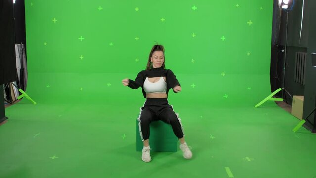 A young woman dancing on a green screen background. Attractive girl making content for social media. Female danser sits on box. Chroma key