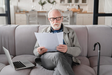 Old elderly senior caucasian man grandfather checking pension documents, loan, e-banking, doing paperwork with laptop, mortgage, paying domestic bills at home