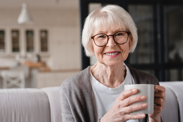 Comfort relaxed senior old elderly woman grandmother drinking hot beverage tea coffee at home looking at camera in the living room. Pension concept.