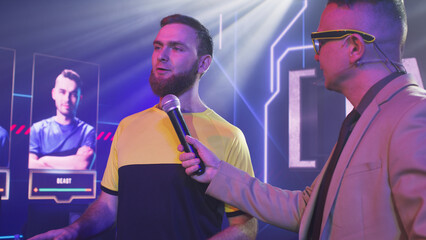 Adult man host with microphone asking questions to bearded gamer esportsman and announcing start of...
