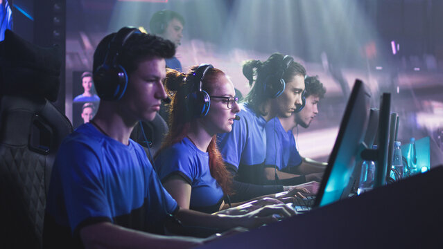 Young and successful men and women professional gamers with coach playing video game on computers against monitor with match broadcast during esports tournament