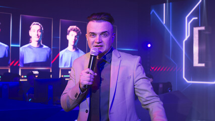 Host with microphone gesticulating and looking at camera while standing between tables with teams...