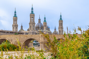 Fototapeta na wymiar Medieval cityscape with ancient stone bridge over yellow water river and Basilica in Zaragoza, Spain. Puente de Piedra, Cathedral-Basilica of Our Lady of the Pillar and Ebro river on an autumn day