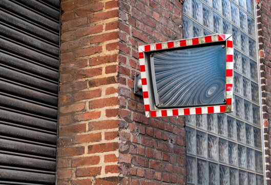 Dented old traffic mirror made of polished metal with red and white chequered frame on the red brick facade of a ruinded factory building in Germany. Rolling gate and glass building blocks.